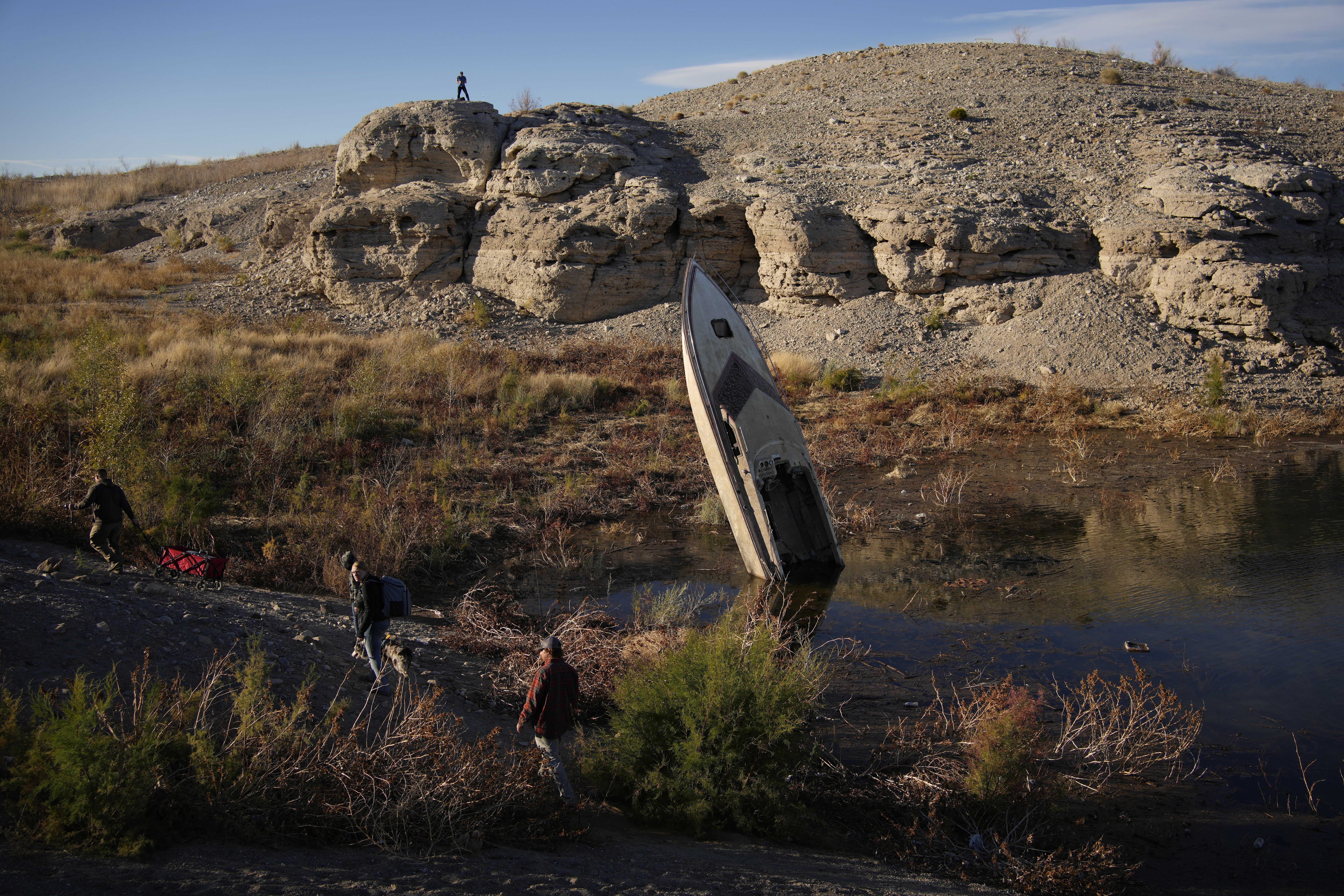 People walk by a formerly sunken boat standing upright into the air with its stern buried in the mud along the shoreline of Lake Mead.
